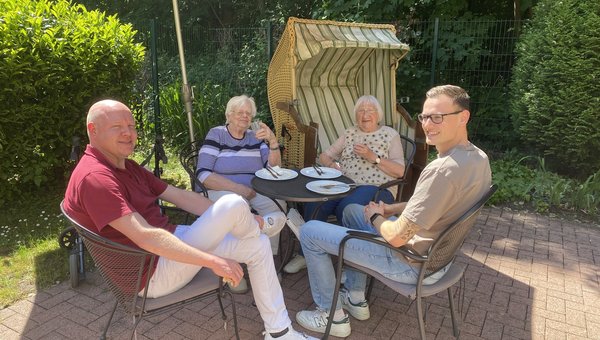 Zwei Seniorinnen sitzen mit zwei Pflegekräften draußen am Tisch, alle lächeln freundlich.