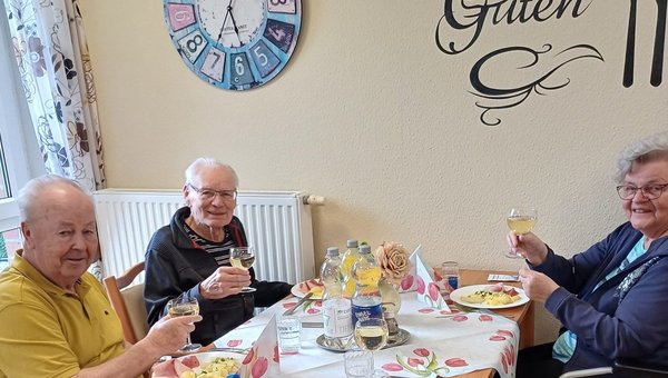 Auf dem Bild sieht man drei Senioren, die beim Essen an einem Tisch sitzen. Sie prosten freundlich mit ihren Gläsern Wein in die Kamera
