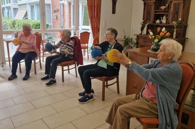 Mehrere Seniorinnen sitzen in einem Halbkreis auf Stühlen beim Rehasport und halten einen Ball mit ausgestreckten Armen.