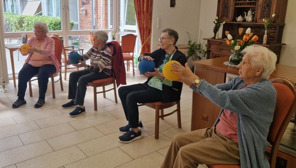 Mehrere Seniorinnen sitzen in einem Halbkreis auf Stühlen beim Rehasport und halten einen Ball mit ausgestreckten Armen.