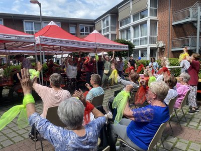 Man blickt über eine Gruppe Senioren, die einen Sitztanz vorführen, ihnen gegenüber steht Publikum, das mitmacht.