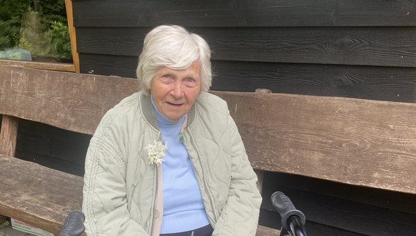 Eine Seniorin sitzt auf einer Holzbank und lächelt in die Kamera.