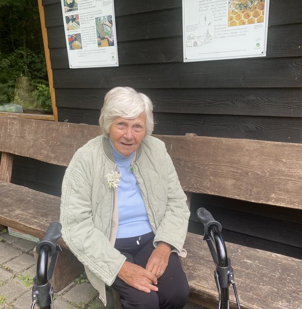 Eine Seniorin sitzt auf einer Holzbank und lächelt in die Kamera. 
