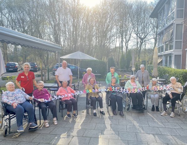 Mehrere Senioren sitzen nebeneinander auf Stühlen, hinter ihnen stehen weitere Senioren. Gemeinsam halten sie eine bunte, selbstgebastelte Menschenkette