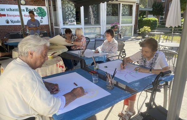 Man sieht mehrere Seniorinnen auf einer Terrasse an Tischen sitzen. Sie haben alle ein großes Blatt weißes Papier vor sich. Alle schauen konzentriert auf das Blatt und malen