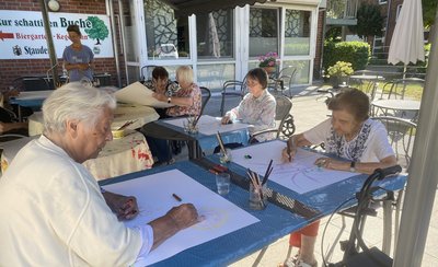 Man sieht mehrere Seniorinnen auf einer Terrasse an Tischen sitzen. Sie haben alle ein großes Blatt weißes Papier vor sich. Alle schauen konzentriert auf das Blatt und malen