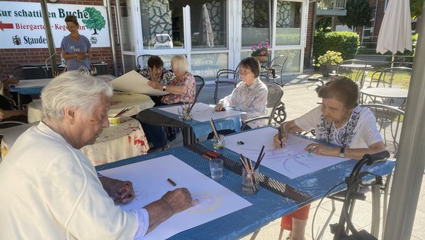 Man sieht mehrere Seniorinnen auf einer Terrasse an Tischen sitzen. Sie haben alle ein großes Blatt weißes Papier vor sich. Alle schauen konzentriert auf das Blatt und malen