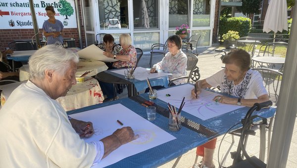Man sieht mehrere Seniorinnen auf einer Terrasse an Tischen sitzen. Sie haben alle ein großes Blatt weißes Papier vor sich. Alle schauen konzentriert auf das Blatt und malen