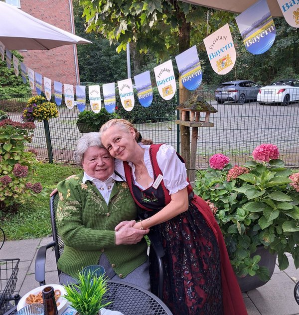 Zwei Frauen in bayerischer Tracht sitzen Arm in Arm, umgeben von bayerischer Dekoration.