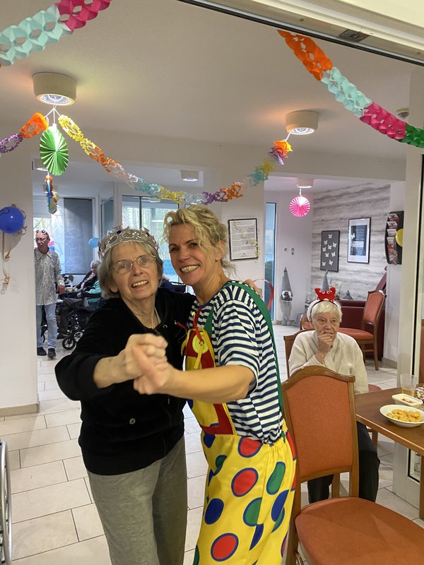 Eine Frau im Clownskostüm tanzt mit einer Seniorin mit Krone, beide lachen in die Kamera.