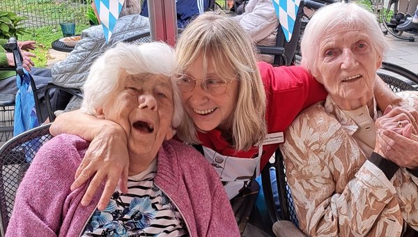 Eine Frau hält zwei Seniorinnen im Arm. Alle lachen herzlich.