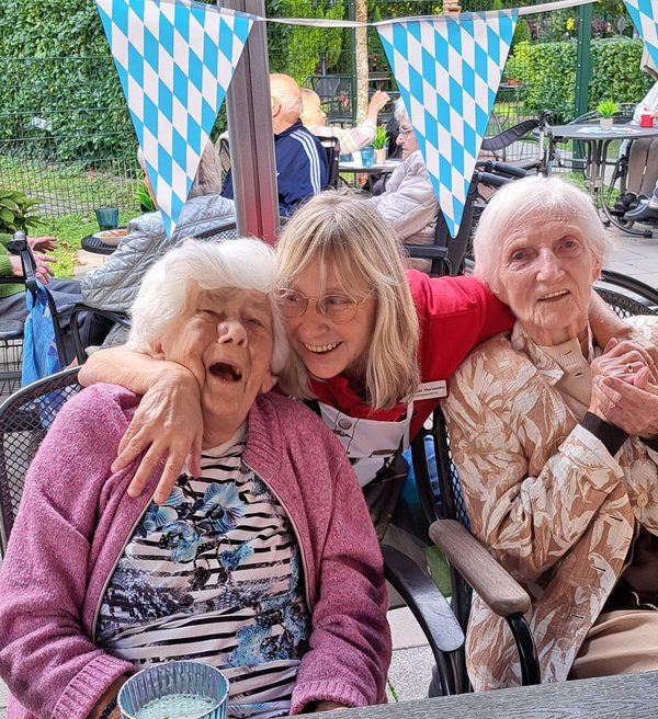 Eine Frau hält zwei Seniorinnen im Arm. Alle lachen herzlich.