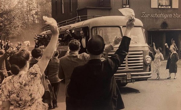 Man sieht einen Bus, aus dem Menschen jubeln. Vor dem Bus stehen Leute, die mit Taschentüchern winken. Im Hintergrund ist eine Gaststätte mit dem Namen "Zur schattigen Buche"
