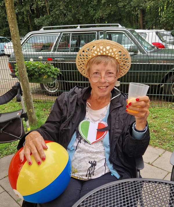 Eine Frau mit Strohhut und einem Italien-Herz-Anhänger um den Hals hält einen Wasserball in der einen und ein Getränk in der anderen Hand. Sie lacht fröhlich in die Kamera.