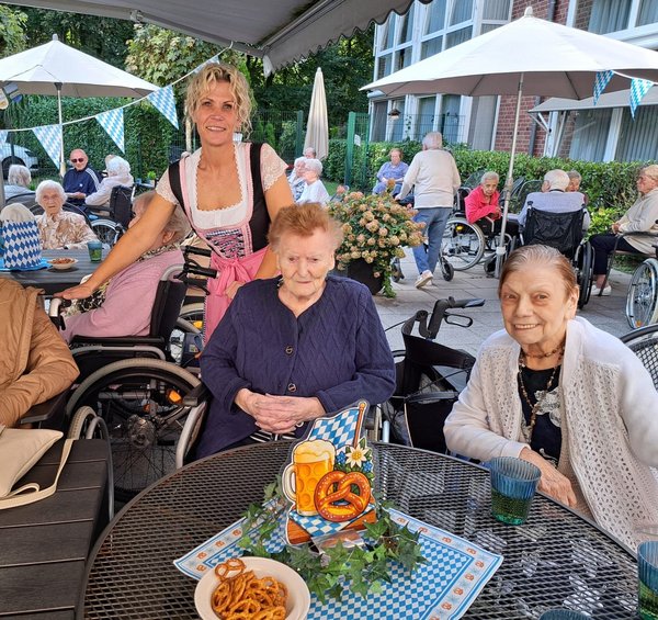 Eine Gruppe Seniorinnen sitzt auf einer bayerisch geschmückten Terrasse, dahinter steht eine Frau im Dirndl.