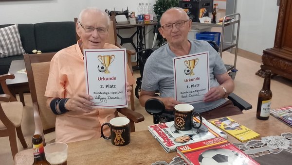 Zwei Herren sitzen an einem Tisch und zeigen ihre Urkunden. Vor ihnen stehen Fußball-Dekorationen. Sie freuen sich über ihren Gewinn beim Bundesliga-Tippspiel.