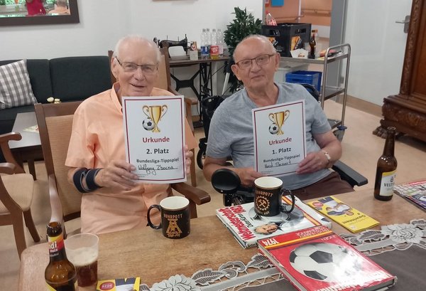 Zwei Herren sitzen an einem Tisch und zeigen ihre Urkunden. Vor ihnen stehen Fußball-Dekorationen. Sie freuen sich über ihren Gewinn beim Bundesliga-Tippspiel.