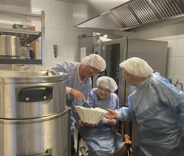 Ein Mann zeigt zwei Seniorinnen in einer Großküche einen großen Kaffeefilter. Alle tragen Kittel und Hauben.