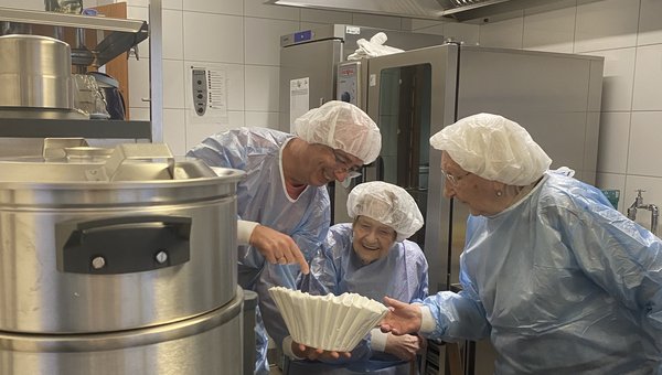 Ein Mann zeigt zwei Seniorinnen in einer Großküche einen großen Kaffeefilter. Alle tragen Kittel und Hauben.