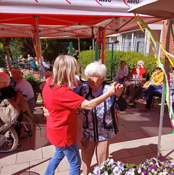 Zwei Frauen tanzen im Vordergrund, im Hintergrund sitzen weitere Menschen unter mit bunten Bändern geschmückten Pavillons.