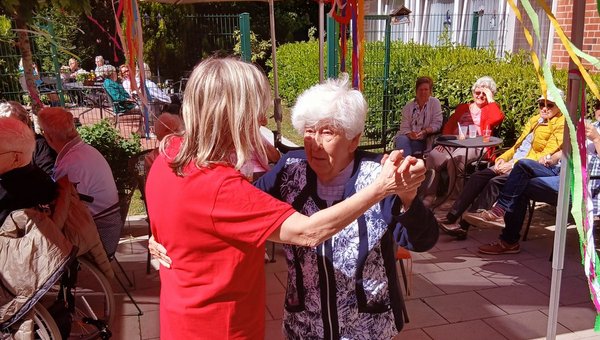 Zwei Frauen tanzen im Vordergrund, im Hintergrund sitzen weitere Menschen unter mit bunten Bändern geschmückten Pavillons.