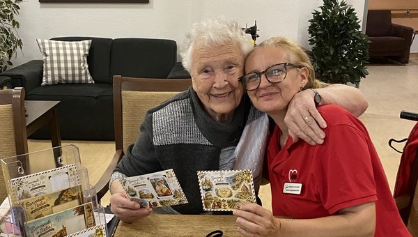 Zwei Frauen sitzen an einem Tisch, auf dem mehrere Postkarten liegen. Sie haben sich im Arm und lachen in die Kamera.