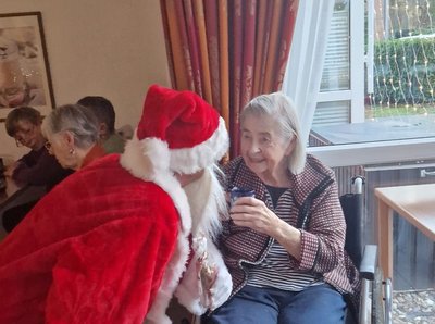 Ein Mann im Nikolauskostüm beugt sich zu einer Seniorin hinunter. Sie lächelt ihn an und hält dabei eine Tasse Kakao in der Hand.