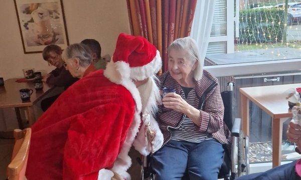 Ein Mann im Nikolauskostüm beugt sich zu einer Seniorin hinunter. Sie lächelt ihn an und hält dabei eine Tasse Kakao in der Hand.