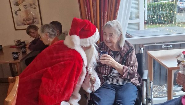 Ein Mann im Nikolauskostüm beugt sich zu einer Seniorin hinunter. Sie lächelt ihn an und hält dabei eine Tasse Kakao in der Hand.