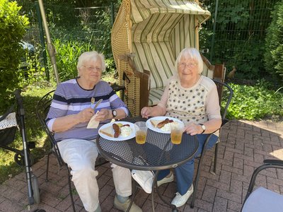 Zwei Seniorinnen sitzen bei schönem Wetter zum Mittagessen auf der Terrasse. Vor ihnen stehen Teller mit Grillwürstchen und Kartoffelsalat.