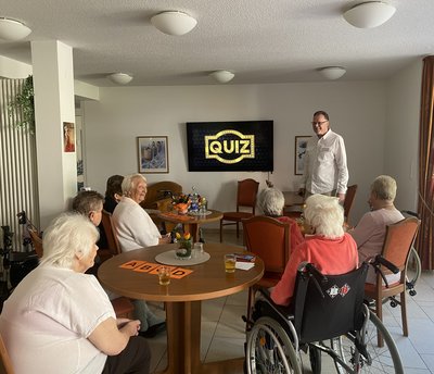 Auf dem Bild sieht man eine Gruppe von Senioren, die auf einen Bildschirm schauen, auf dem eine Quiz steht. Neben dem Bildschirm steht der Moderator.