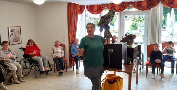 Auf dem Bild sieht man eine Frau, die eine Uhr auf dem Arm hält. Im Hintergrund sieht man Kinder und Senioren sitzen