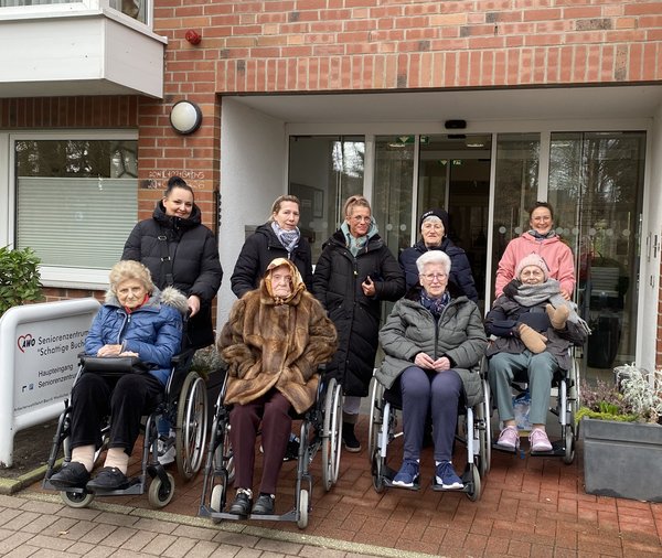 Mehrere Senior*innen sitzen in Rollstühlen, dahinter stehen Mitarbeitende. Alle tragen warme Jacken, Schals und Mützen und lächeln in die Kamera. 