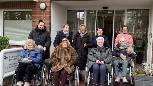 Mehrere Senior*innen sitzen in Rollstühlen, dahinter stehen Mitarbeitende. Alle tragen warme Jacken, Schals und Mützen und lächeln in die Kamera. 