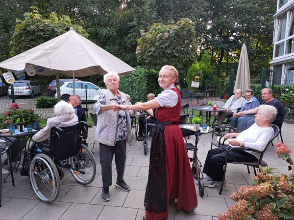 Eine Frau im Dirndl tanzt mit einer Seniorin auf der Terrasse.