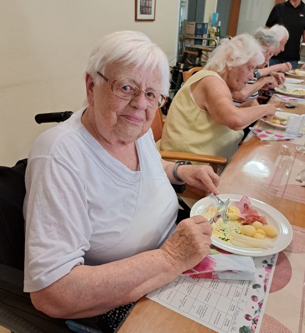 Eine Seniorin blickt freundlich in die Kamera. Vor ihr steht ein Teller mit Spargel, Schinken, Kartoffeln und Sauce Hollandaise.