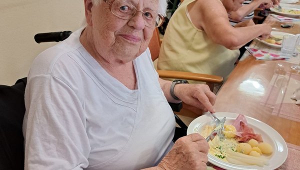 Eine Seniorin blickt freundlich in die Kamera. Vor ihr steht ein Teller mit Spargel, Schinken, Kartoffeln und Sauce Hollandaise.