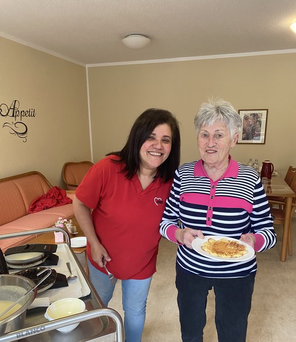Zwei Frauen lächeln in die Kamera, eine hält einen Teller mit einer Waffel, neben ihnen sind Waffeleisen und der Teig zu sehen.