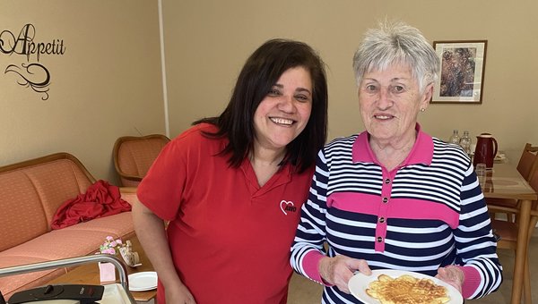 Zwei Frauen lächeln in die Kamera, eine hält einen Teller mit einer Waffel, neben ihnen sind Waffeleisen und der Teig zu sehen.