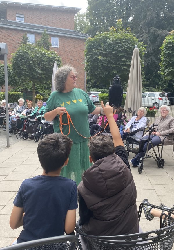 Eine Frau hält einen Raben auf dem Arm. Im Vordergrund sitzen zwei Kinder, eines zeigt auf. Im Hintergrund sitzen Seniorinnen.