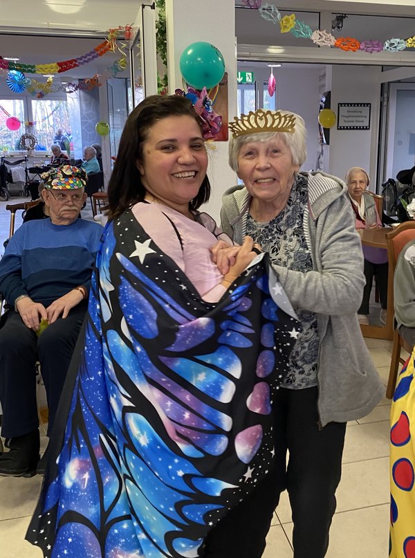 Eine Frau im Schmetterlingskostüm tanzt mit einer Seniorin, die eine Krone trägt, beide lachen in die Kamera