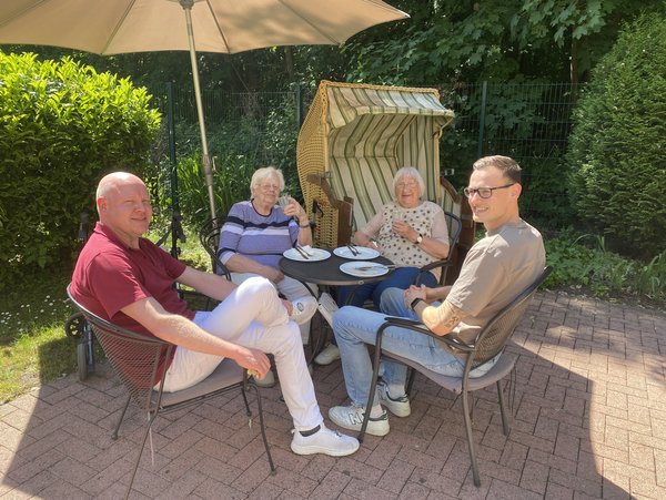 Zwei Seniorinnen sitzen mit zwei Pflegekräften draußen am Tisch, alle lächeln freundlich.