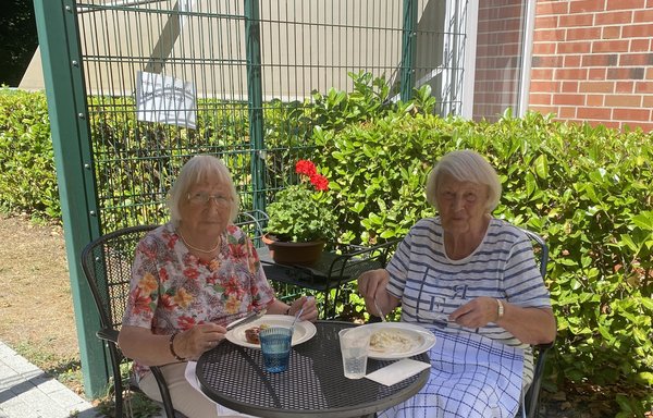 Zwei Seniorinnen sitzen auf einer Terrasse und essen Würstchen mit Kartoffelsalat.