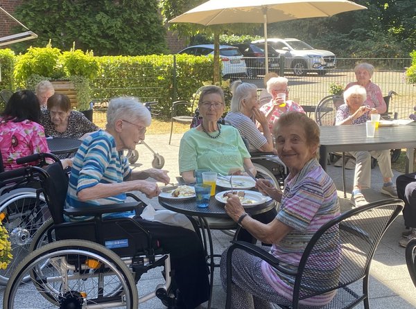Mehrere Seniorinnen sitzen gemeinsam auf einer Terrasse und essen Grillwurst mit Kartoffelsalat. Eine der Seniorinnen schaut lächelnd über ihre Schulter in die Kamera.t.