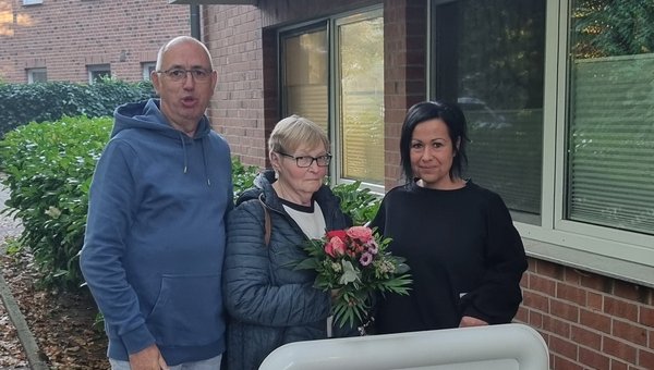 Auf dem Bild sieht man drei Personen vor einem Seniorenzentrum stehen. Die Frau in der Mitte hält einen Blumenstrauß.