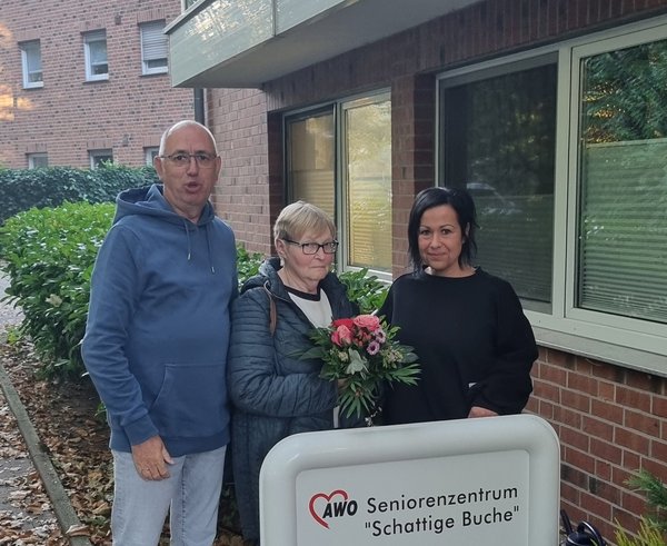 Auf dem Bild sieht man drei Personen vor einem Seniorenzentrum stehen. Die Frau in der Mitte hält einen Blumenstrauß.