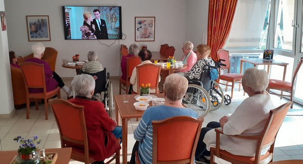 Eine Gruppe von Bewohnern sitzt mit dem Rücken zu uns und blickt gebannt auf den Bildschirm, während sie einen gemütlichen Kino-Nachmittag genießen.