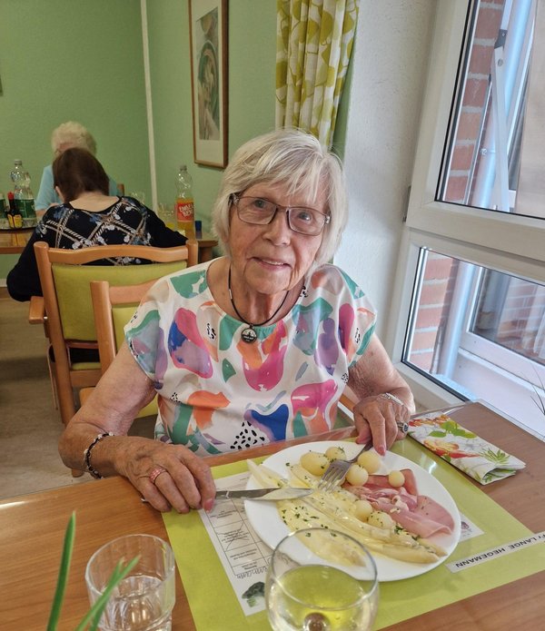 Eine Seniorin blickt freundlich in die Kamera. Vor ihr steht ein Teller mit Spargel, Schinken, Kartoffeln und Sauce Hollandaise.