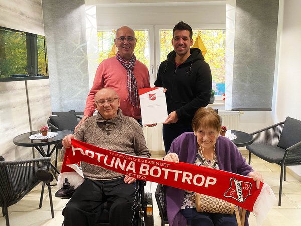 In der vorderen Reihe sitzen zwei Senioren, die einen Fortuna-Bottrop-Schal halten. Dahinter stehen zwei Männer, die gemeinsam eine Mappe von Fortuna Bottrop hochhalten.