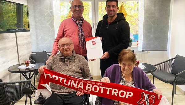 In der vorderen Reihe sitzen zwei Senioren, die einen Fortuna-Bottrop-Schal halten. Dahinter stehen zwei Männer, die gemeinsam eine Mappe von Fortuna Bottrop hochhalten.